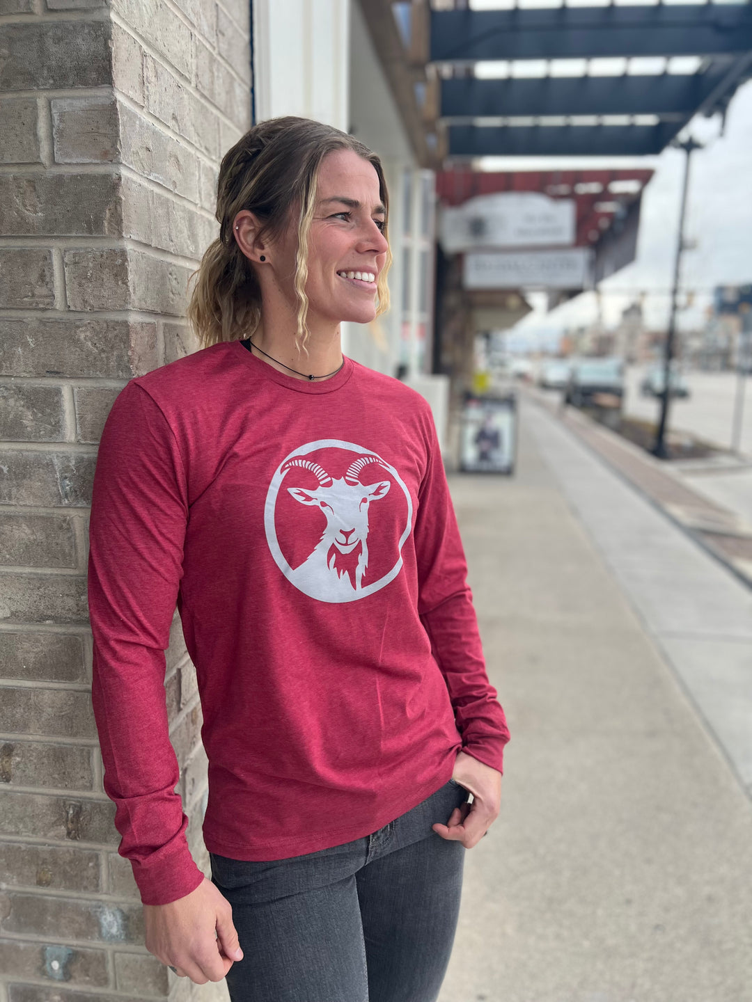 Stylish woman wearing a long-sleeve Heather Cardinal shirt with a white goat logo centered on the chest, standing outside against a brick wall, representing the adventurous spirit of Timpanogos Hiking Co.