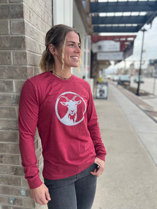 Stylish woman wearing a long-sleeve Heather Cardinal shirt with a white goat logo centered on the chest, standing outside against a brick wall, representing the adventurous spirit of Timpanogos Hiking Co.