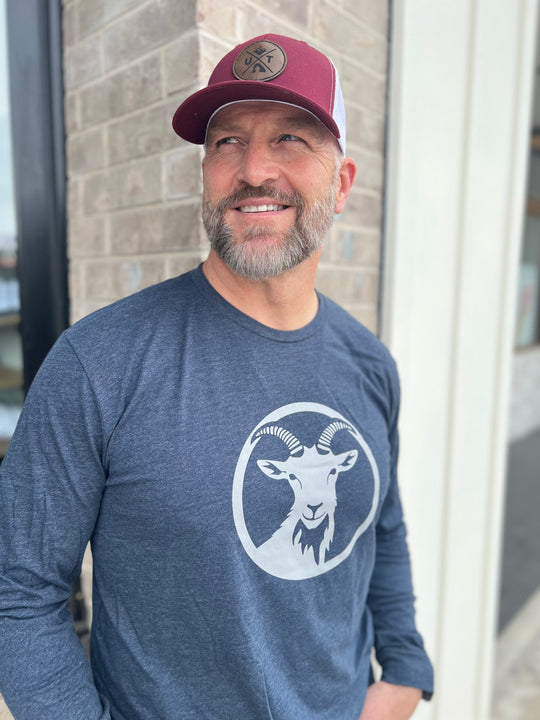 Man wearing a long-sleeve Heather Navy shirt with a white goat graphic in a circle, paired with a maroon and white mesh cap, standing outside against a brick wall, showcasing Timpanogos Hiking Co.'s outdoor apparel.