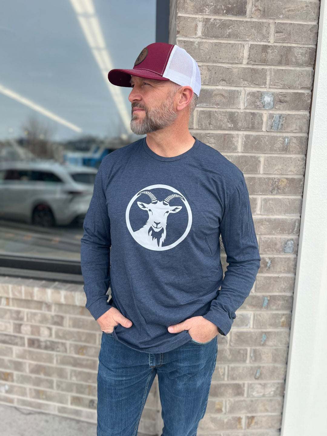Man wearing a long-sleeve Heather Navy shirt with a white goat graphic in a circle, paired with a maroon and white mesh cap, standing outside against a brick wall, showcasing Timpanogos Hiking Co.'s outdoor apparel.