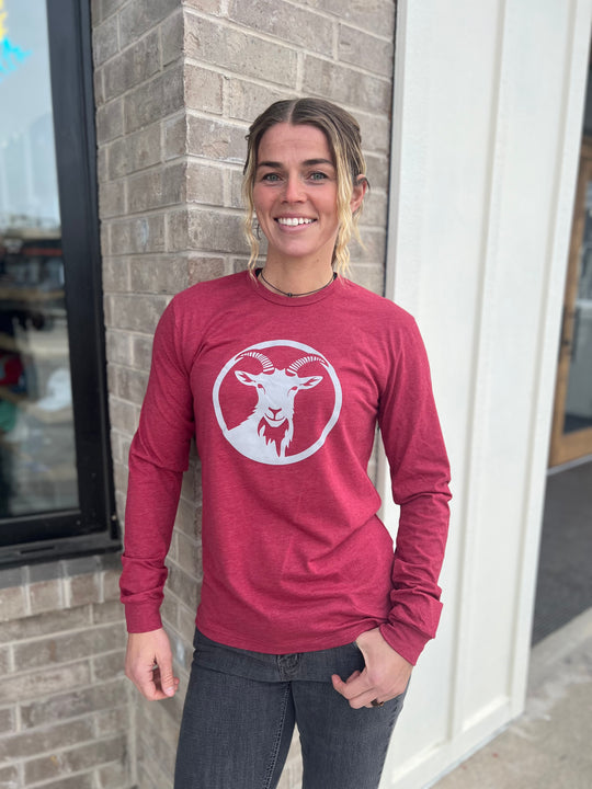 Stylish woman wearing a long-sleeve Heather Cardinal shirt with a white goat logo centered on the chest, standing outside against a brick wall, representing the adventurous spirit of Timpanogos Hiking Co.