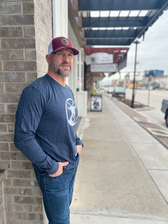Man wearing a long-sleeve Heather Navy shirt with a white goat graphic in a circle, paired with a maroon and white mesh cap, standing outside against a brick wall, showcasing Timpanogos Hiking Co.'s outdoor apparel.