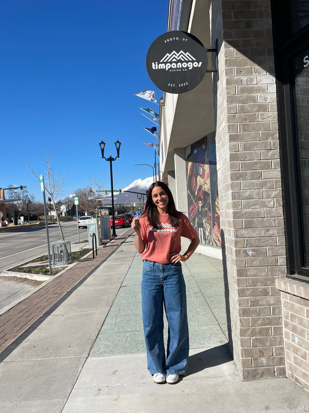 Woman standing outside the Timpanogos Hiking Co. storefront in Provo, Utah, wearing a faded red graphic tee with the Timpanogos logo and wide-leg blue jeans. She smiles while holding a round sticker, with snow-capped Mount Timpanogos visible in the background under a clear blue sky.