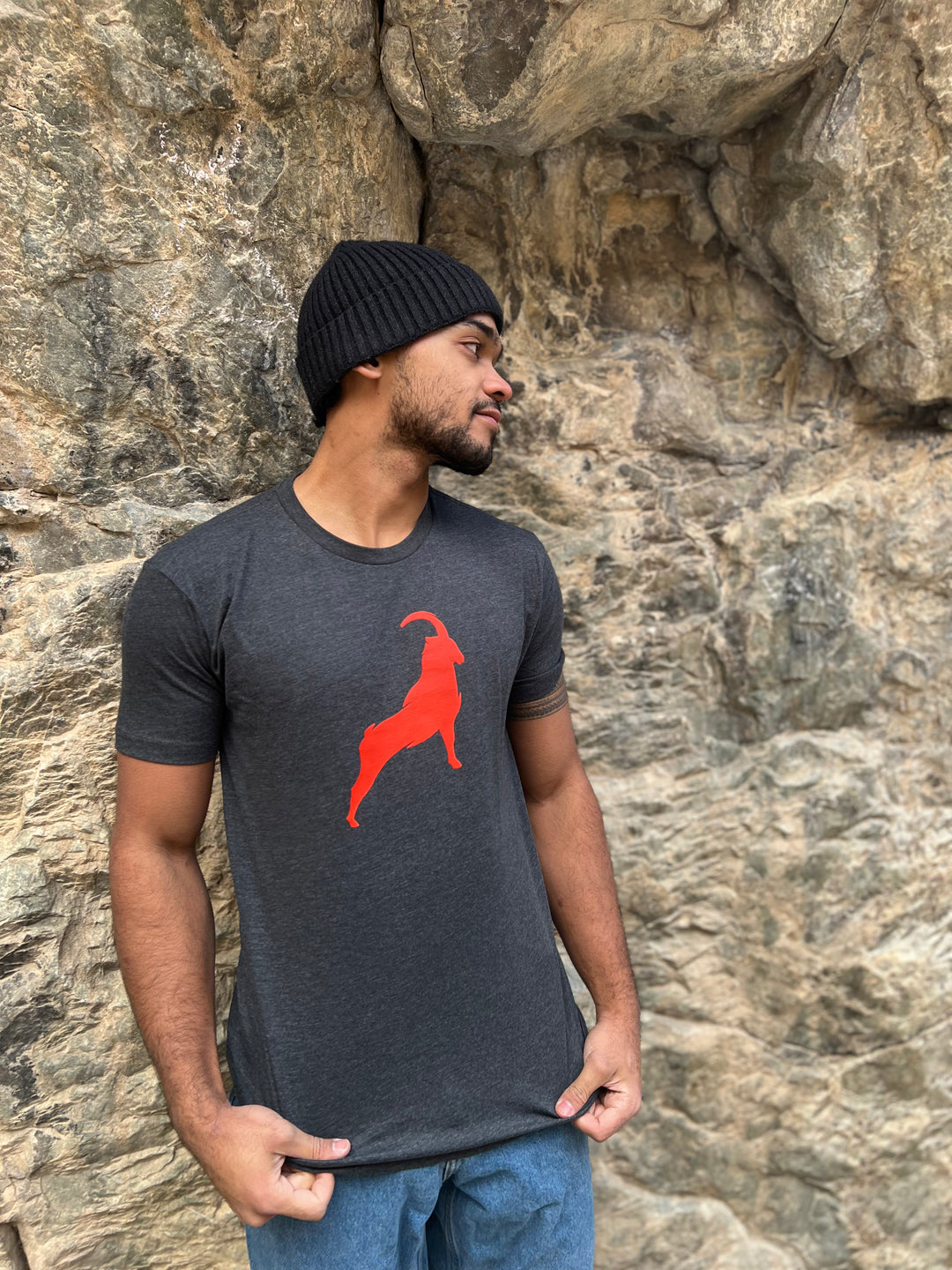 Man wearing a Heather Charcoal T-shirt with a bold orange goat graphic, standing against a rocky backdrop, highlighting the adventurous outdoor style of Timpanogos Hiking Co.