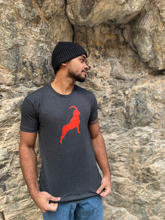 Man wearing a Heather Charcoal T-shirt with a bold orange goat graphic, standing against a rocky backdrop, highlighting the adventurous outdoor style of Timpanogos Hiking Co.