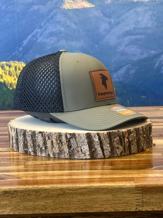 Heather Military Green trucker hat with a brown leather Timpanogos Hiking Co. goat logo patch, displayed on a wooden stump with the Wasatch Mountains in the background.