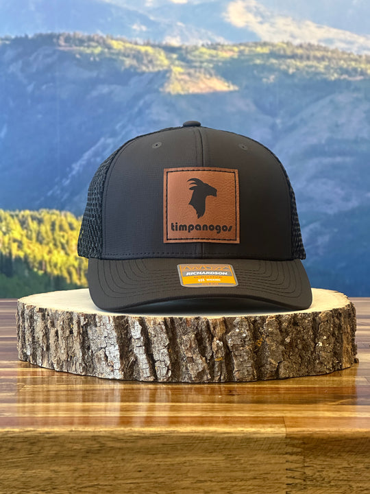 Heather Black trucker hat featuring a brown leather Timpanogos goat patch, resting on a wooden stump with a backdrop of mountains.