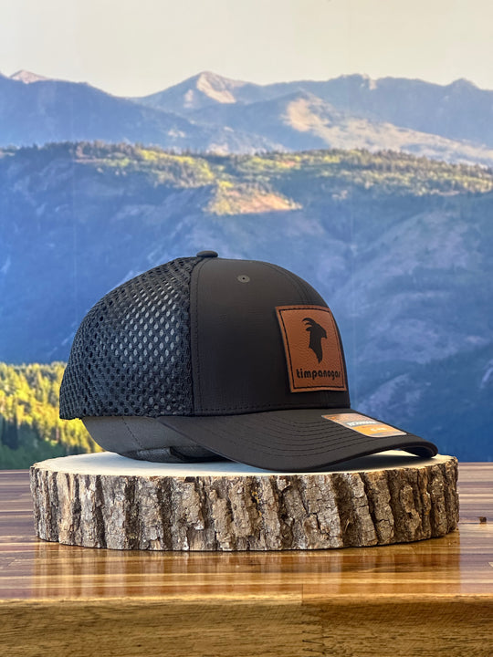 Timpanogos Hiking Co. Heather Black trucker hat featuring a leather goat patch, placed on a wooden surface with a scenic mountain view in the background.