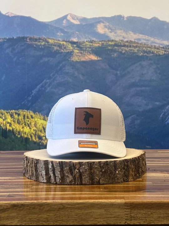 White trucker hat with a brown leather patch featuring the Timpanogos Hiking Co. goat logo, displayed on a wooden surface with the Wasatch Mountains in the background.