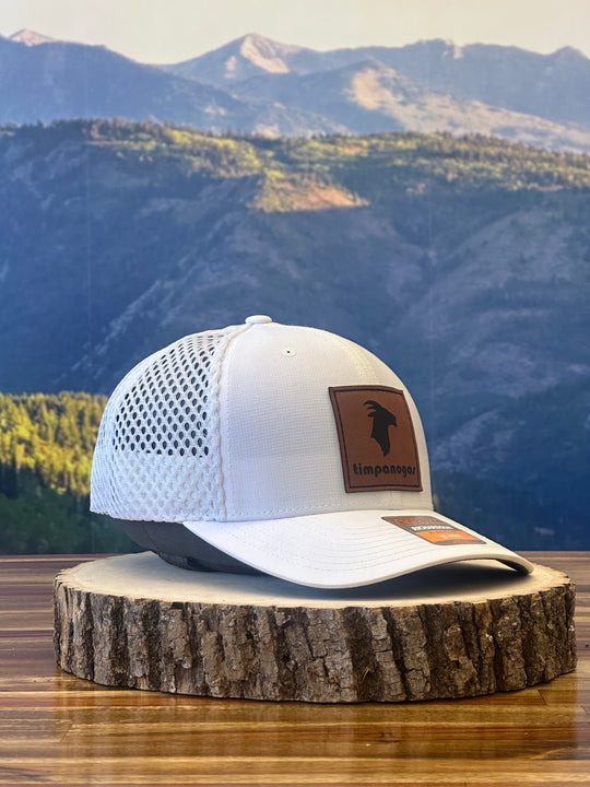 White trucker hat featuring a brown leather patch with a goat graphic and "Timpanogos" text, placed on a rustic wood slab with a mountain landscape in the background