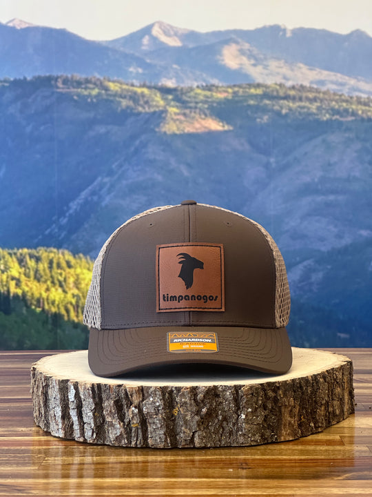 Timpanogos Hiking Co. Heather Espresso trucker hat featuring a leather goat patch, placed on a wooden surface with a scenic mountain view in the background.