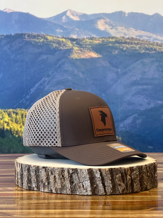 Heather Espresso trucker hat with a brown leather patch featuring the Timpanogos Hiking Co. logo and a goat graphic, displayed on a wooden surface with a backdrop of mountains.