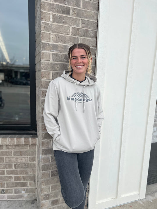 Woman wearing a Heather Cream hoodie with the Timpanogos Hiking Co. mountain logo, standing outdoors against a brick wall with a storefront in the background, showcasing the casual outdoor style of Timpanogos Hiking Co.