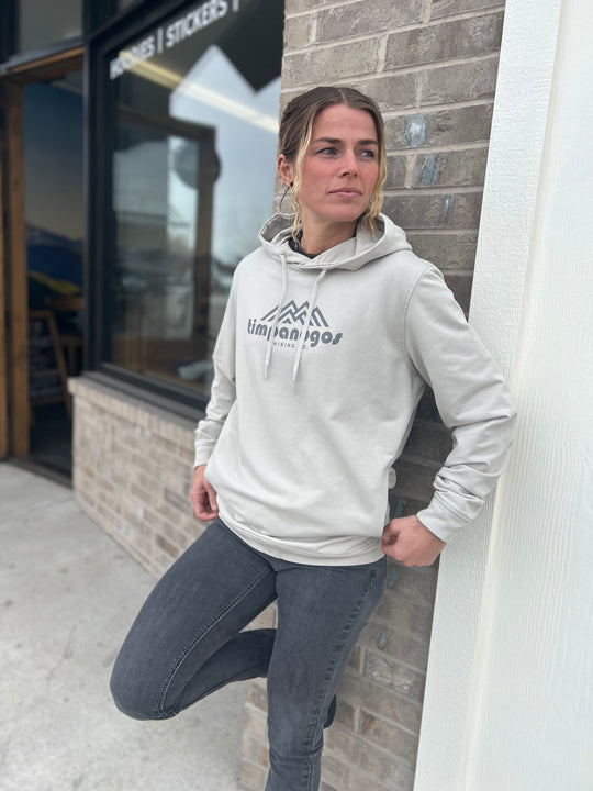 Woman wearing a Heather Cream hoodie with the Timpanogos Hiking Co. mountain logo, standing outdoors against a brick wall with a storefront in the background, showcasing the casual outdoor style of Timpanogos Hiking Co.