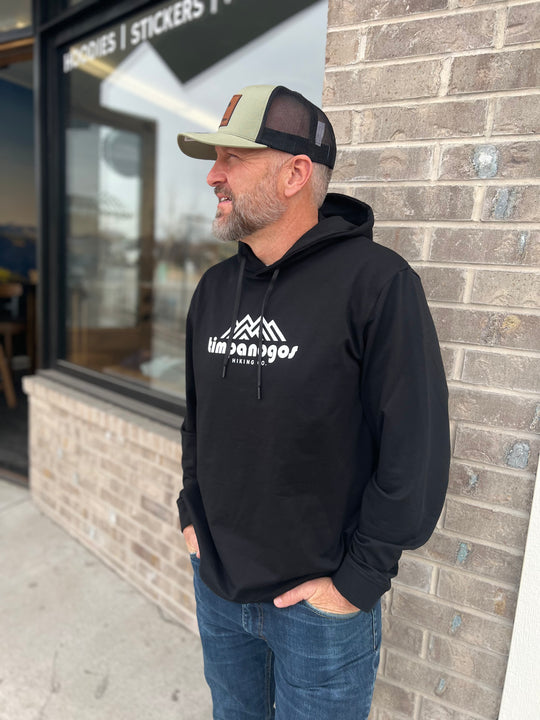 Man wearing a black hoodie with the Timpanogos Hiking Co. logo, paired with a green and black trucker hat, standing outdoors against a brick wall, showcasing the rugged outdoor style of Timpanogos Hiking Co.