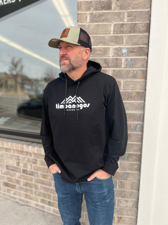 Man wearing a black hoodie with the Timpanogos Hiking Co. logo, paired with a green and black trucker hat, standing outdoors against a brick wall, showcasing the rugged outdoor style of Timpanogos Hiking Co.