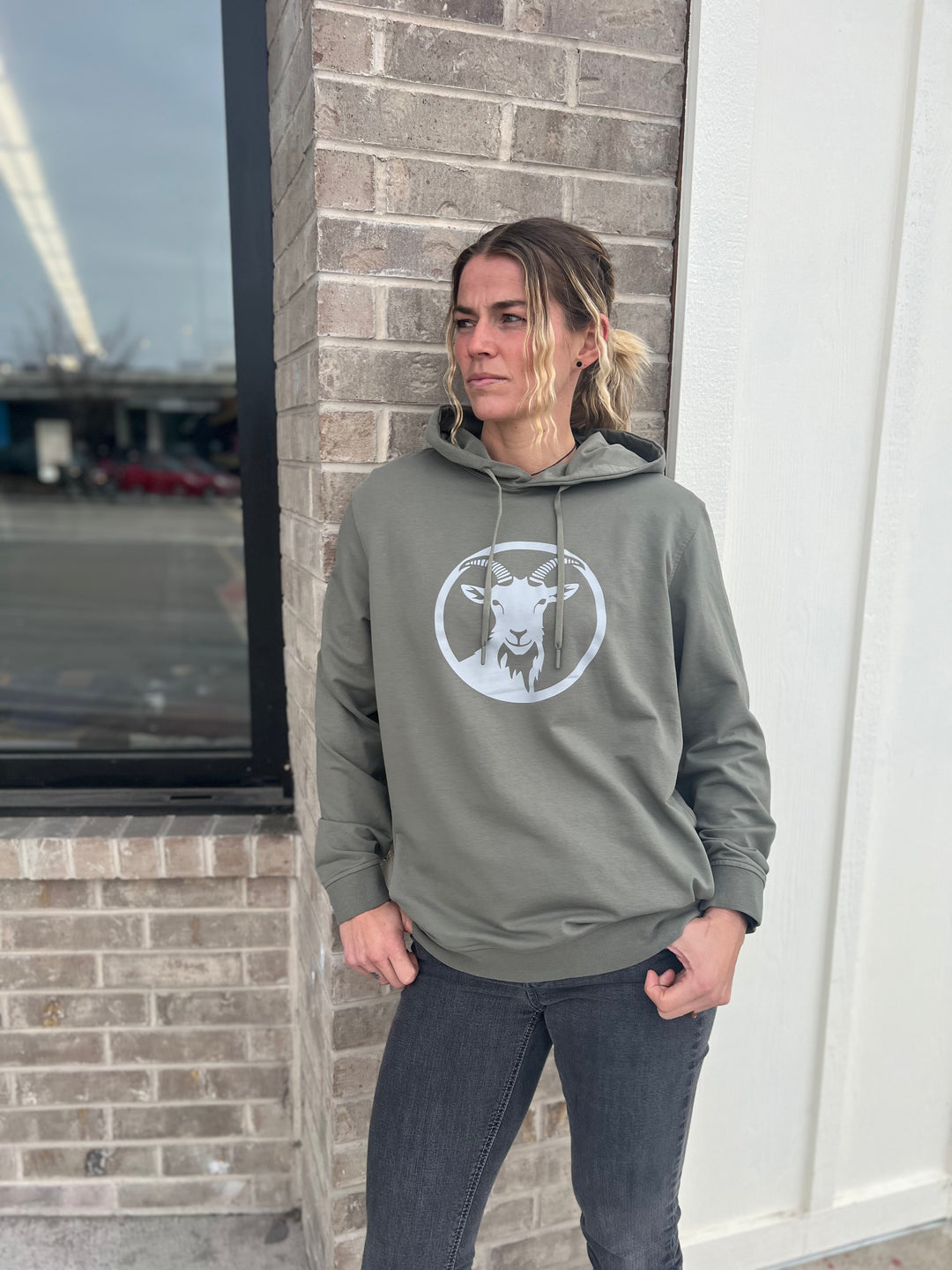Woman wearing a Heather Olive Green hoodie with a white goat graphic in a circle, standing outdoors against a brick wall, representing the adventurous spirit of Timpanogos Hiking Co.