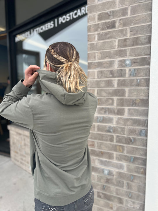 Woman with braided hair pulling up the hood of a Heather Olive Green hoodie, standing outside near a brick wall with a sign in the background, showcasing the casual outdoor style of Timpanogos Hiking Co.