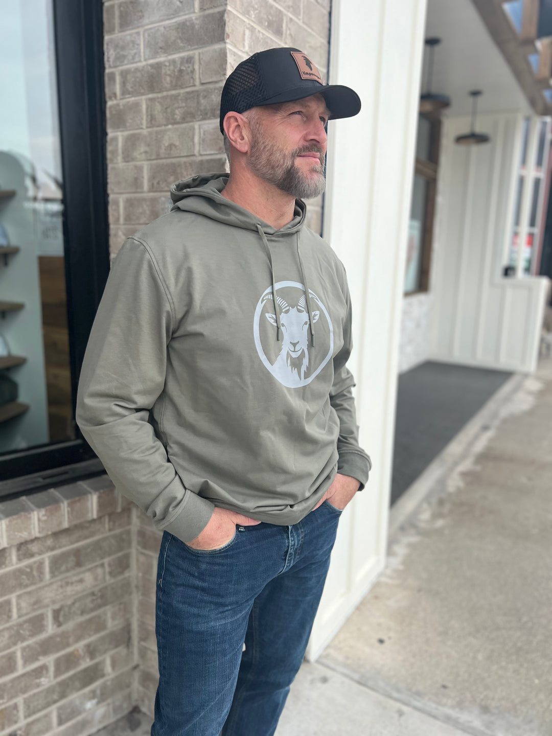 Outdoor enthusiast in a Heather Olive Green hoodie featuring a bold goat logo and circle design, wearing a black trucker hat, standing confidently by a brick building, representing the adventurous spirit of Timpanogos Hiking Co.