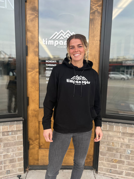 Woman smiling in a black hoodie with the Timpanogos Hiking Co. mountain logo, standing in front of a storefront with a wooden door, showcasing the adventurous outdoor style of Timpanogos Hiking Co.
