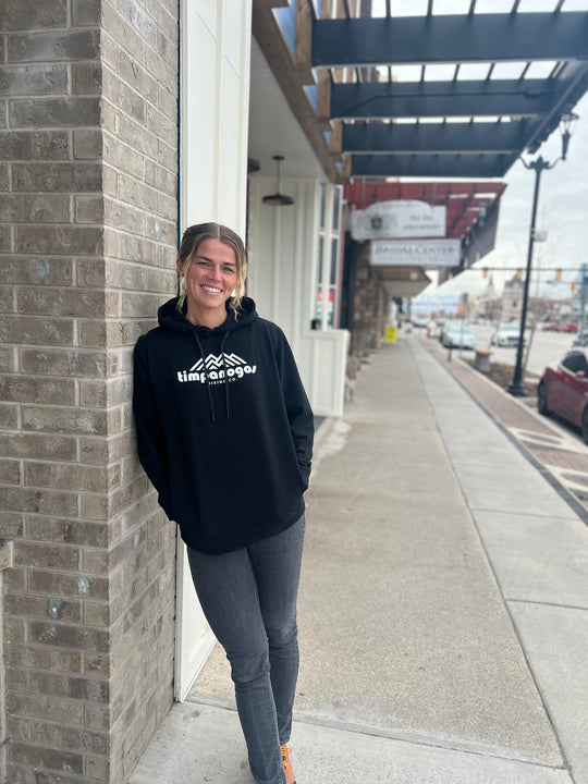 Woman smiling in a black hoodie with the Timpanogos Hiking Co. logo, standing outdoors against a brick wall with a city street backdrop, showcasing the adventurous spirit of Timpanogos Hiking Co.