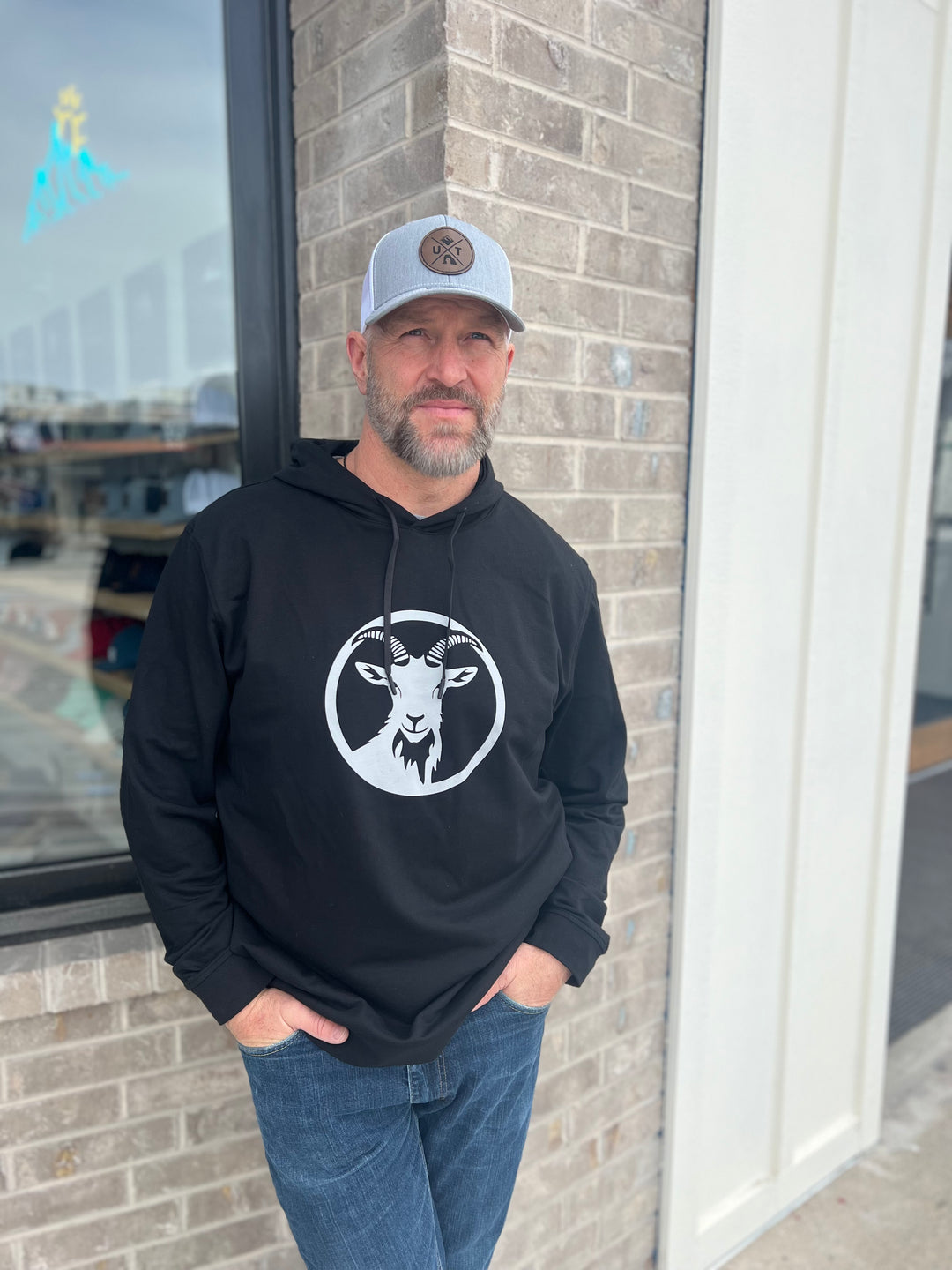 Man wearing a black hoodie with a white goat graphic in a circle, paired with a gray trucker hat, standing outside against a brick wall, representing Timpanogos Hiking Co.'s rugged outdoor apparel.