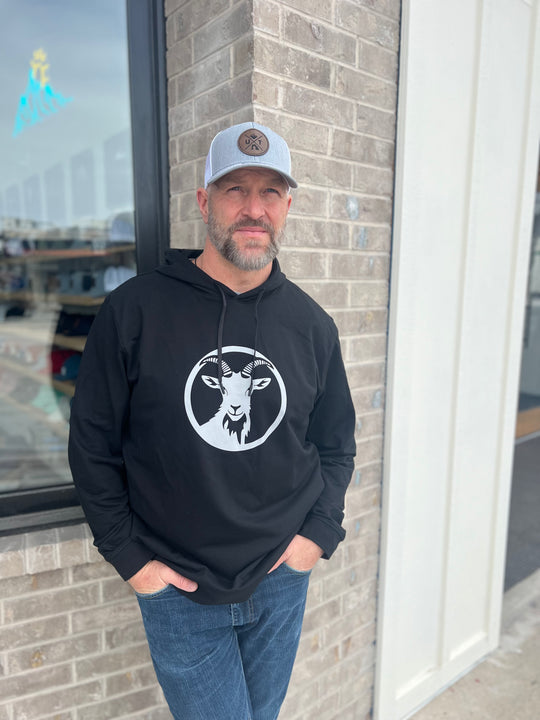 Man wearing a black hoodie with a white goat graphic in a circle, paired with a gray trucker hat, standing outside against a brick wall, representing Timpanogos Hiking Co.'s rugged outdoor apparel.