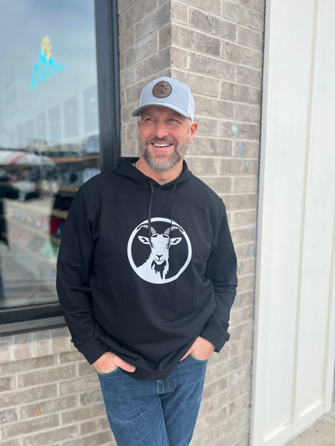 Man wearing a black hoodie with a white goat graphic in a circle, paired with a gray trucker hat, standing outside against a brick wall, representing Timpanogos Hiking Co.'s rugged outdoor apparel.