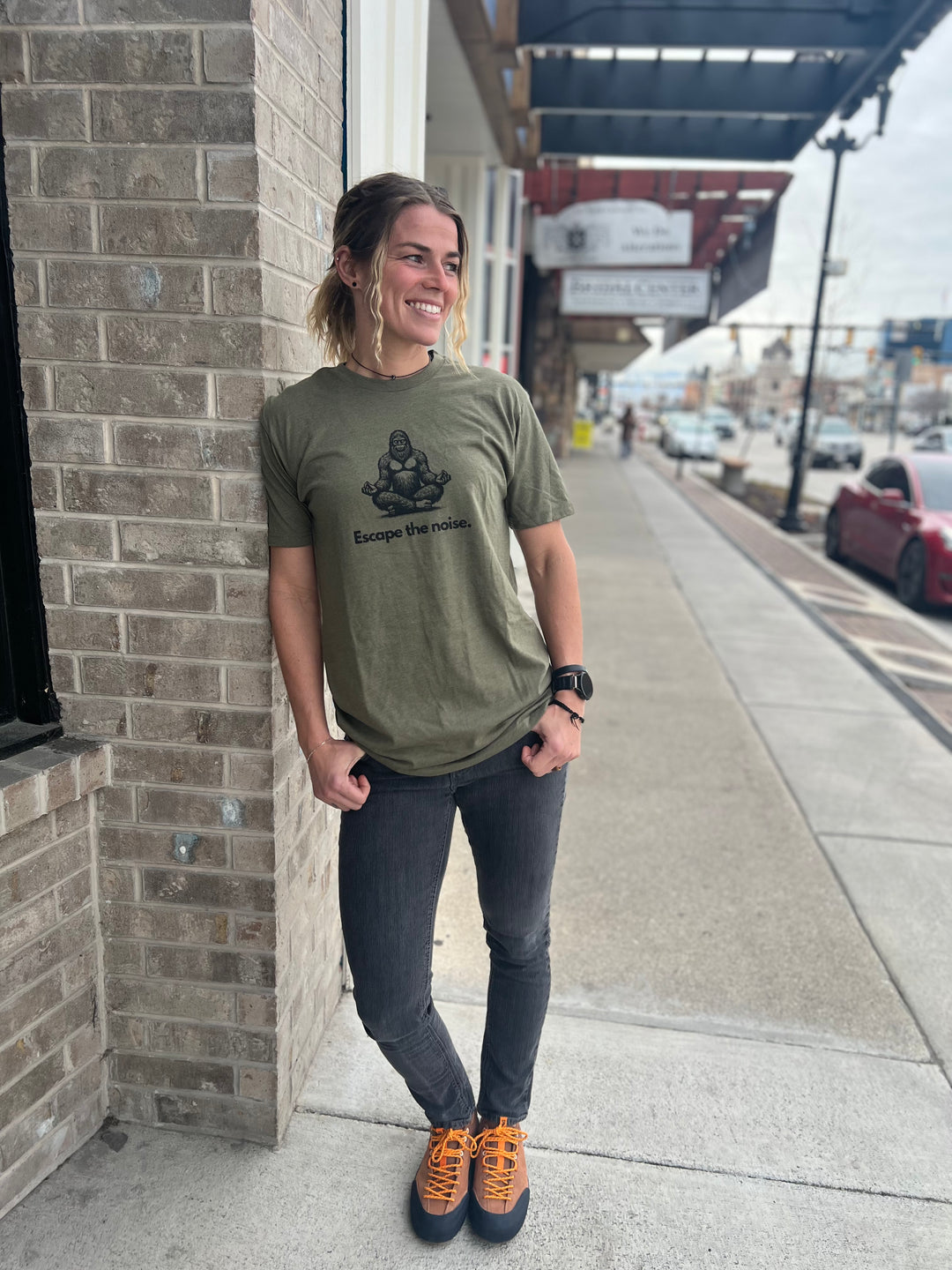 Woman wearing a Heather Military Green T-shirt with a Bigfoot graphic in a meditative pose and the text "Escape the noise," standing outside against a brick wall, showcasing Timpanogos Hiking Co.'s outdoor apparel.