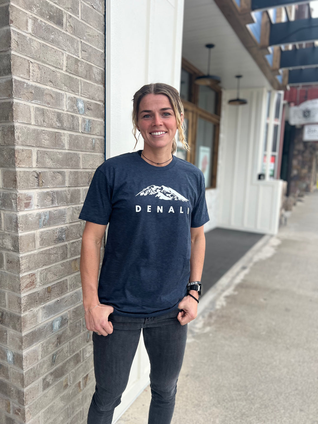 Woman wearing a Heather Navy T-shirt with a white "Denali" mountain graphic, smiling and standing in front of a brick building, representing Timpanogos Hiking Co.'s outdoor apparel.