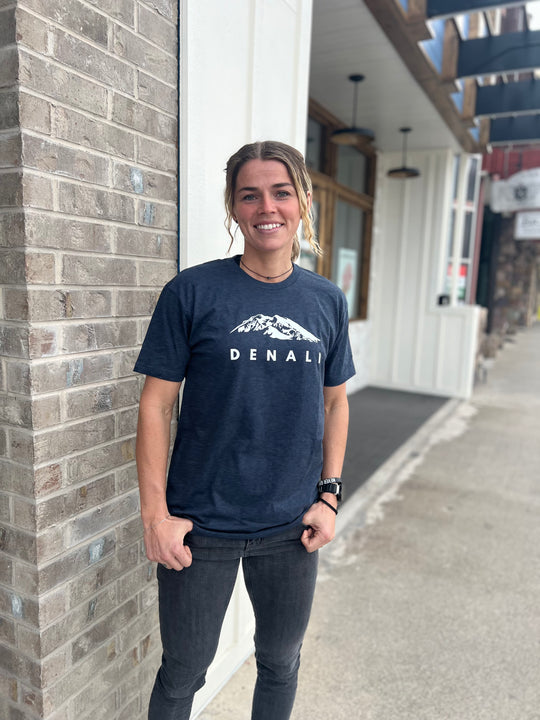 Woman wearing a Heather Navy T-shirt with a white "Denali" mountain graphic, smiling and standing in front of a brick building, representing Timpanogos Hiking Co.'s outdoor apparel.