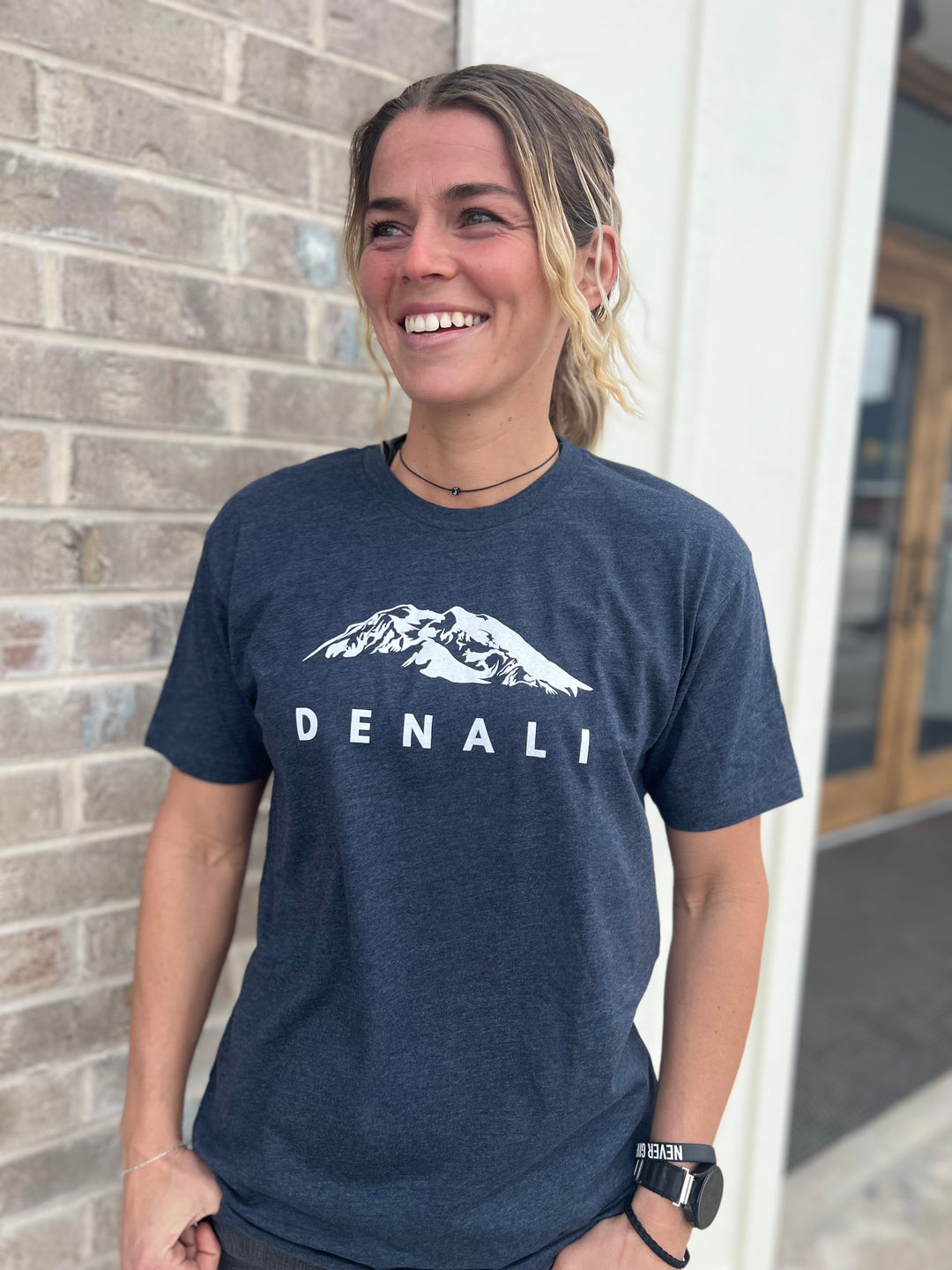 Woman wearing a Heather Navy T-shirt with a white "Denali" mountain graphic, smiling and standing in front of a brick building, representing Timpanogos Hiking Co.'s outdoor apparel.