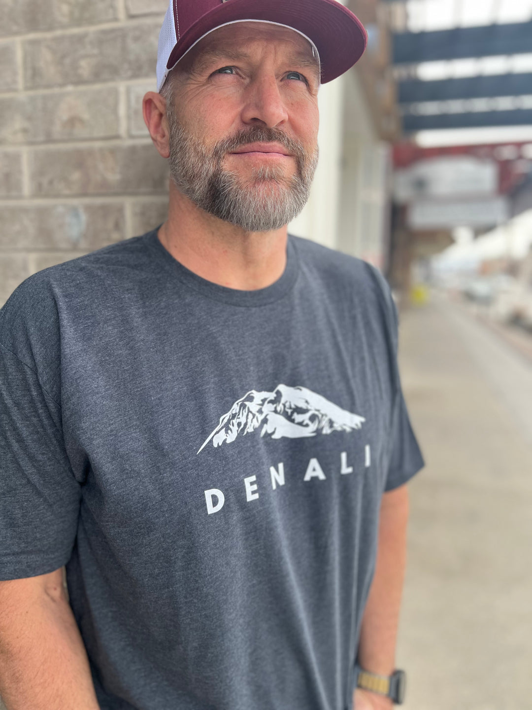 Man wearing a Heather Charcoal T-shirt with a white "Denali" mountain graphic, standing confidently in front of a city street backdrop, complemented by a maroon and white mesh cap, showcasing Timpanogos Hiking Co.'s outdoor apparel.