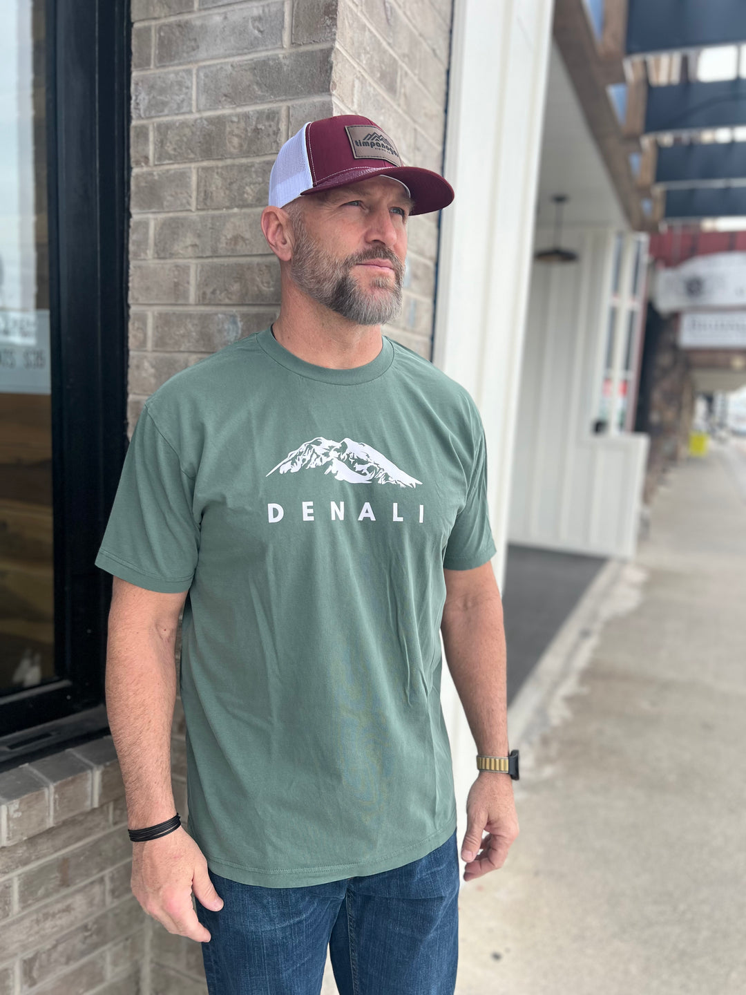 Man wearing a Heather Military Green T-shirt with a white "Denali" mountain graphic, paired with a maroon and white mesh cap, standing outside against a brick wall, representing Timpanogos Hiking Co.'s rugged outdoor spirit.