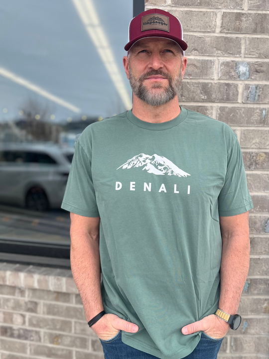 Man wearing a Heather Military Green T-shirt with a white "Denali" mountain graphic, paired with a maroon and white mesh cap, standing outside against a brick wall, representing Timpanogos Hiking Co.'s rugged outdoor spirit.