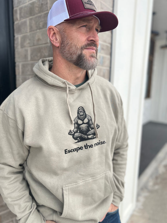 Man wearing a Heather Cream hoodie featuring a graphic of a meditating Bigfoot and the phrase "Escape the noise." Paired with a maroon and white mesh cap, standing outside against a brick wall, representing Timpanogos Hiking Co