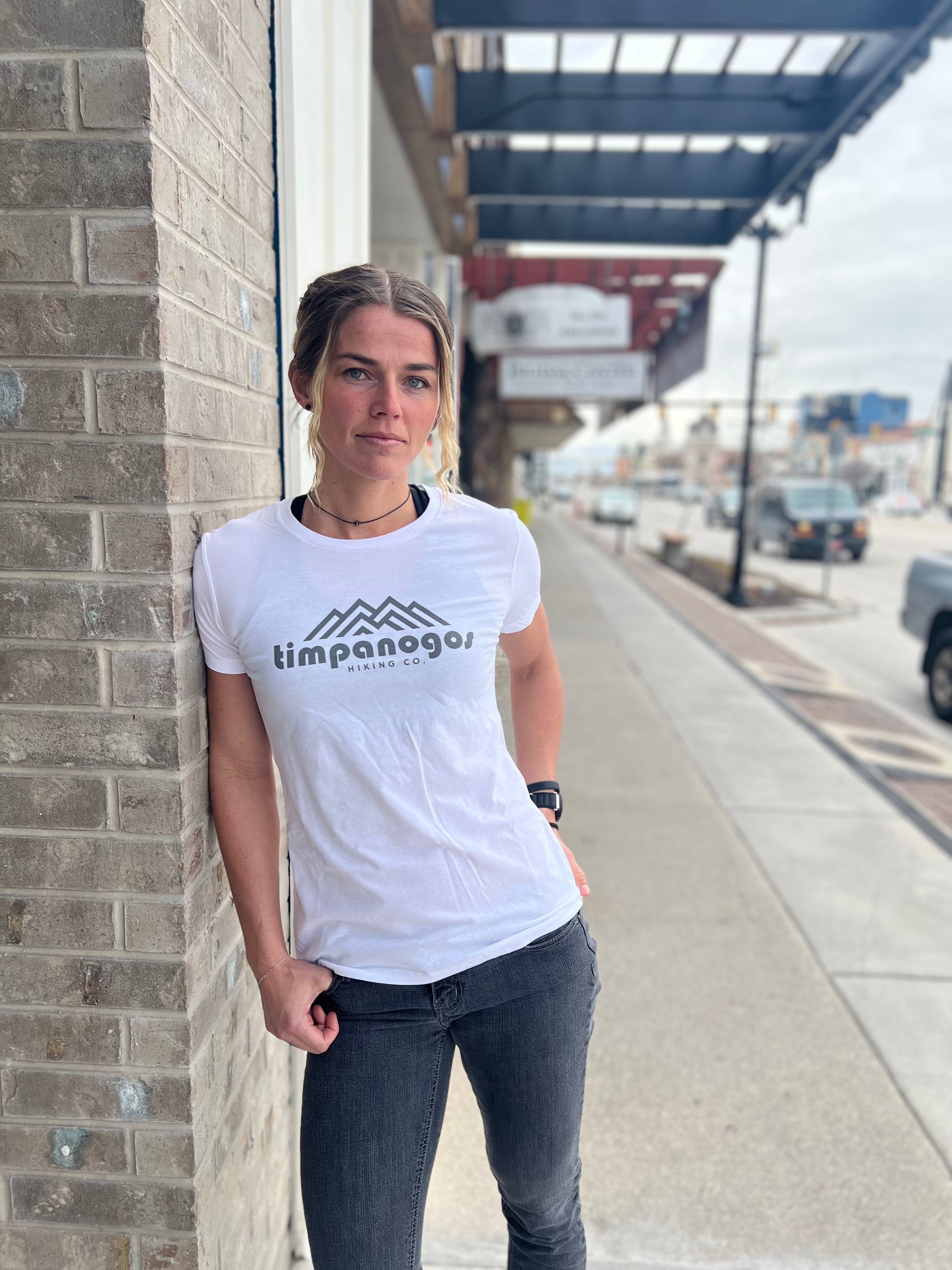 Woman wearing a white T-shirt with the Timpanogos Hiking Co. logo in gray, standing outside against a brick wall with a city street in the background, showcasing the outdoor spirit of Timpanogos Hiking Co.