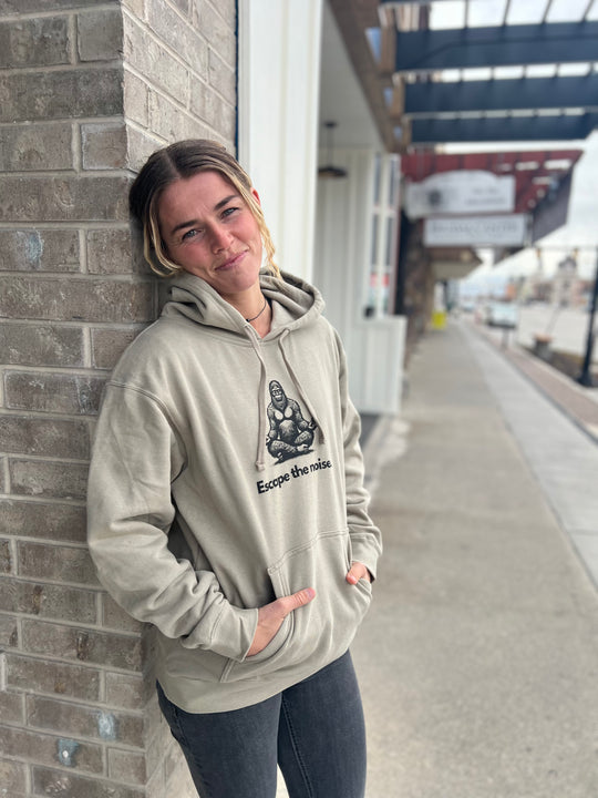 Woman wearing a Heather Cream hoodie featuring a Bigfoot graphic and the "Escape the noise" slogan, standing outside against a brick wall, showcasing Timpanogos Hiking Co.'s adventurous style.