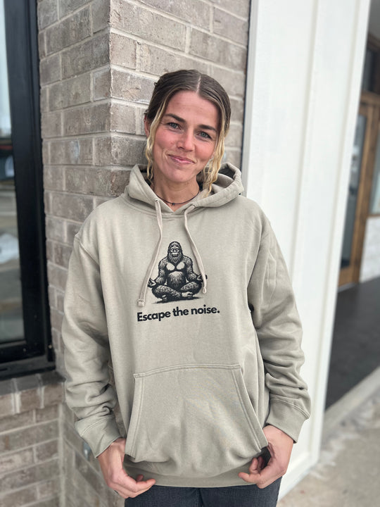 Smiling woman modeling a Heather Cream hoodie with a Bigfoot design and "Escape the noise" message, standing in front of a brick building, highlighting the outdoor spirit of Timpanogos Hiking Co.