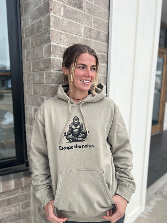 Smiling woman modeling a Heather Cream hoodie with a Bigfoot design and "Escape the noise" message, standing in front of a brick building, highlighting the outdoor spirit of Timpanogos Hiking Co.