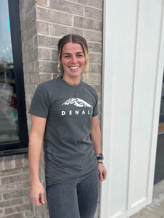 Woman wearing a Heather Gray T-shirt with a white "Denali" mountain graphic, smiling and standing outside against a brick wall, showcasing Timpanogos Hiking Co.'s outdoor apparel.