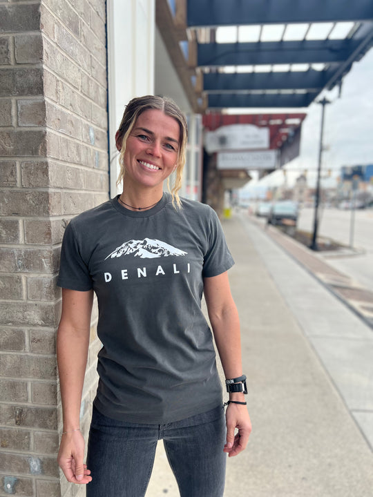 Woman wearing a Heather Gray T-shirt with a white "Denali" mountain graphic, smiling and standing outside against a brick wall, showcasing Timpanogos Hiking Co.'s outdoor apparel.