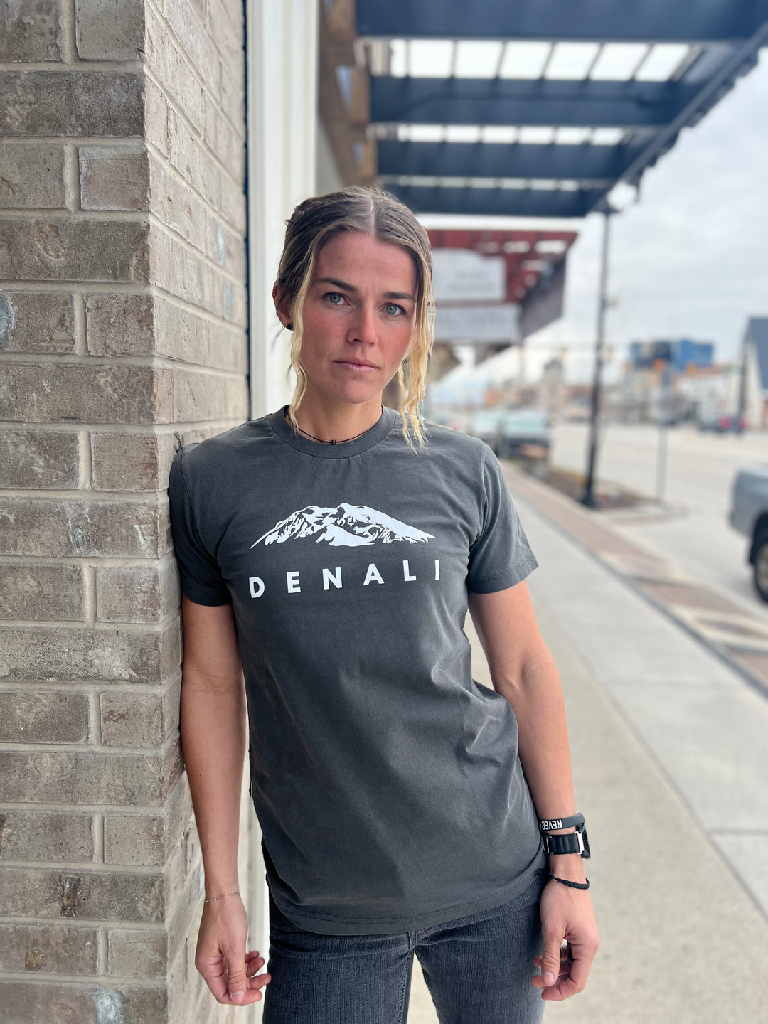 Heather Charcoal t-shirt featuring a white graphic of the Denali mountain range with the word "DENALI" beneath, worn by a woman standing against a brick wall in an urban setting