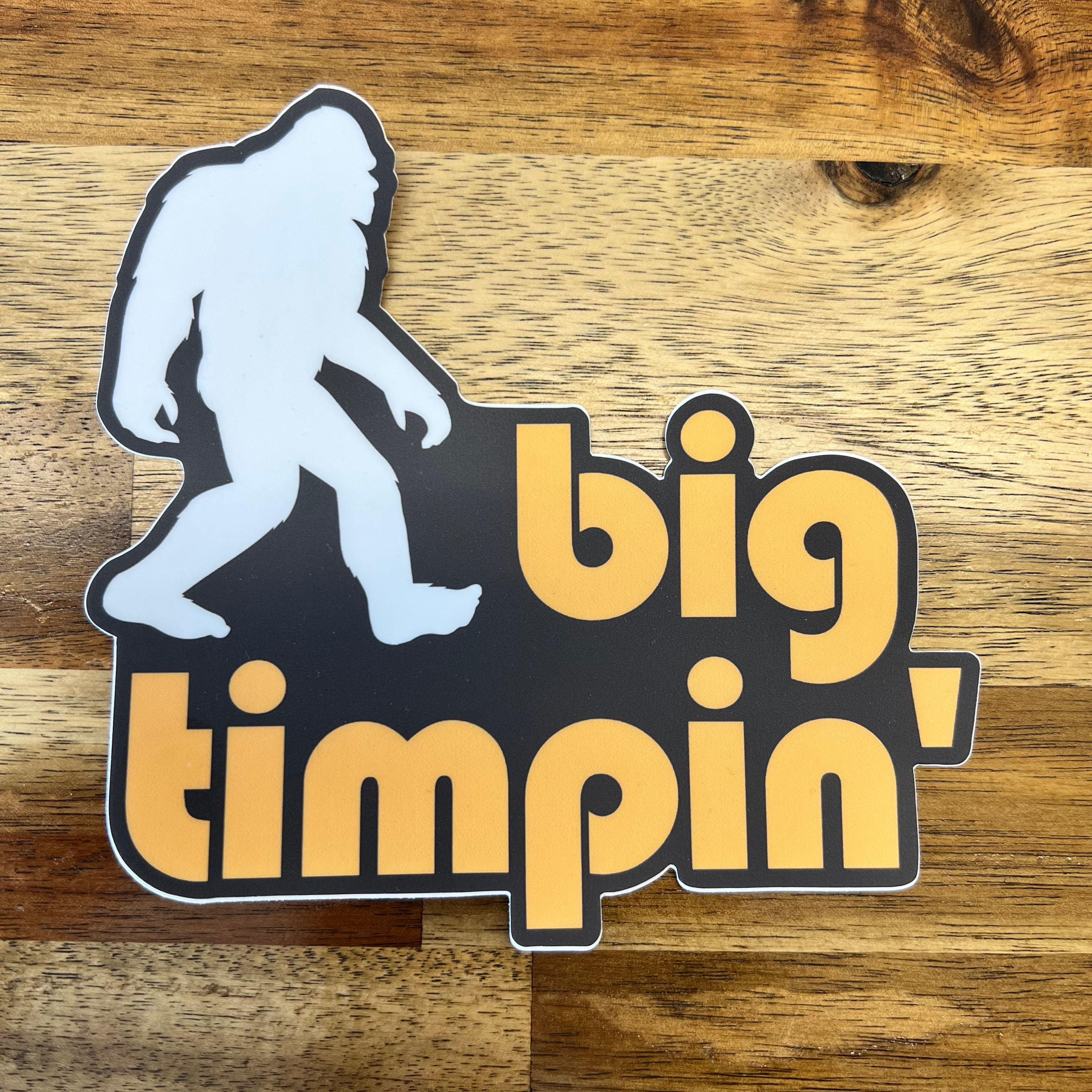Vinyl sticker featuring a white Bigfoot silhouette stepping over bold retro-style yellow-orange text that reads "big timpin'," set against a black background with a white border—perfect for outdoor gear, water bottles, or laptops.