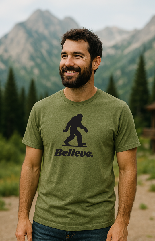 Man wearing an olive green “Believe” Sasquatch graphic t-shirt by Timpanogos Hiking Co., photographed in the Utah mountains — unisex outdoor apparel celebrating mystery and adventure.