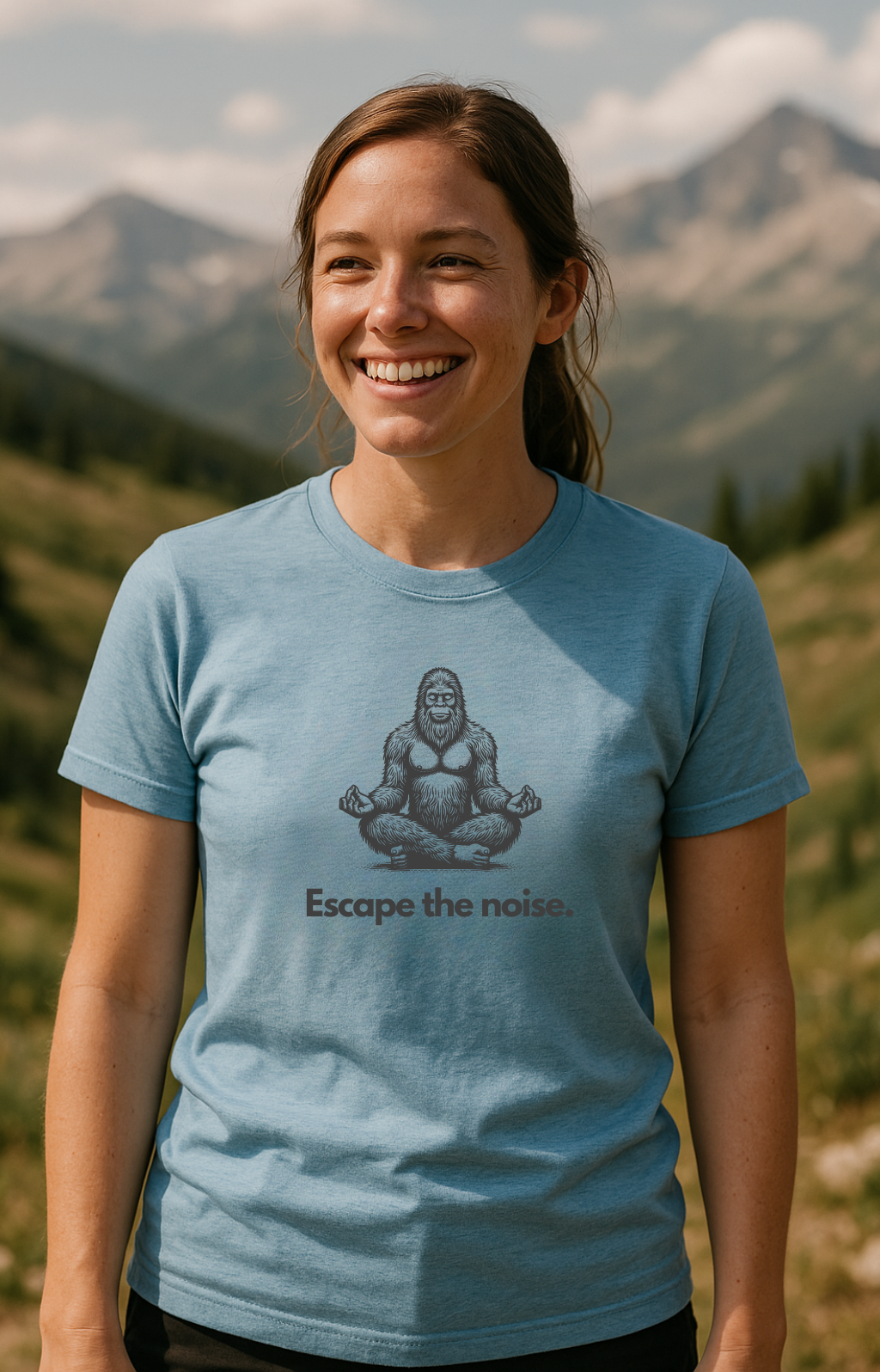 Woman standing in a mountainous outdoor setting wearing a light blue t-shirt. The shirt features an illustration of a meditating Bigfoot and the phrase “Escape the noise.” written below in a clean, modern font. The backdrop includes alpine peaks and scattered pine trees, suggesting a peaceful, remote location.