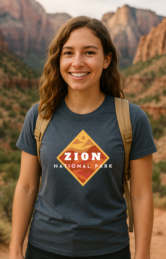 Woman smiling while hiking in Zion National Park, wearing a navy blue graphic tee with a diamond-shaped illustration of canyon layers in warm tones. The design reads “Zion National Park” in bold white text. Towering red rock cliffs and desert greenery appear in the background.