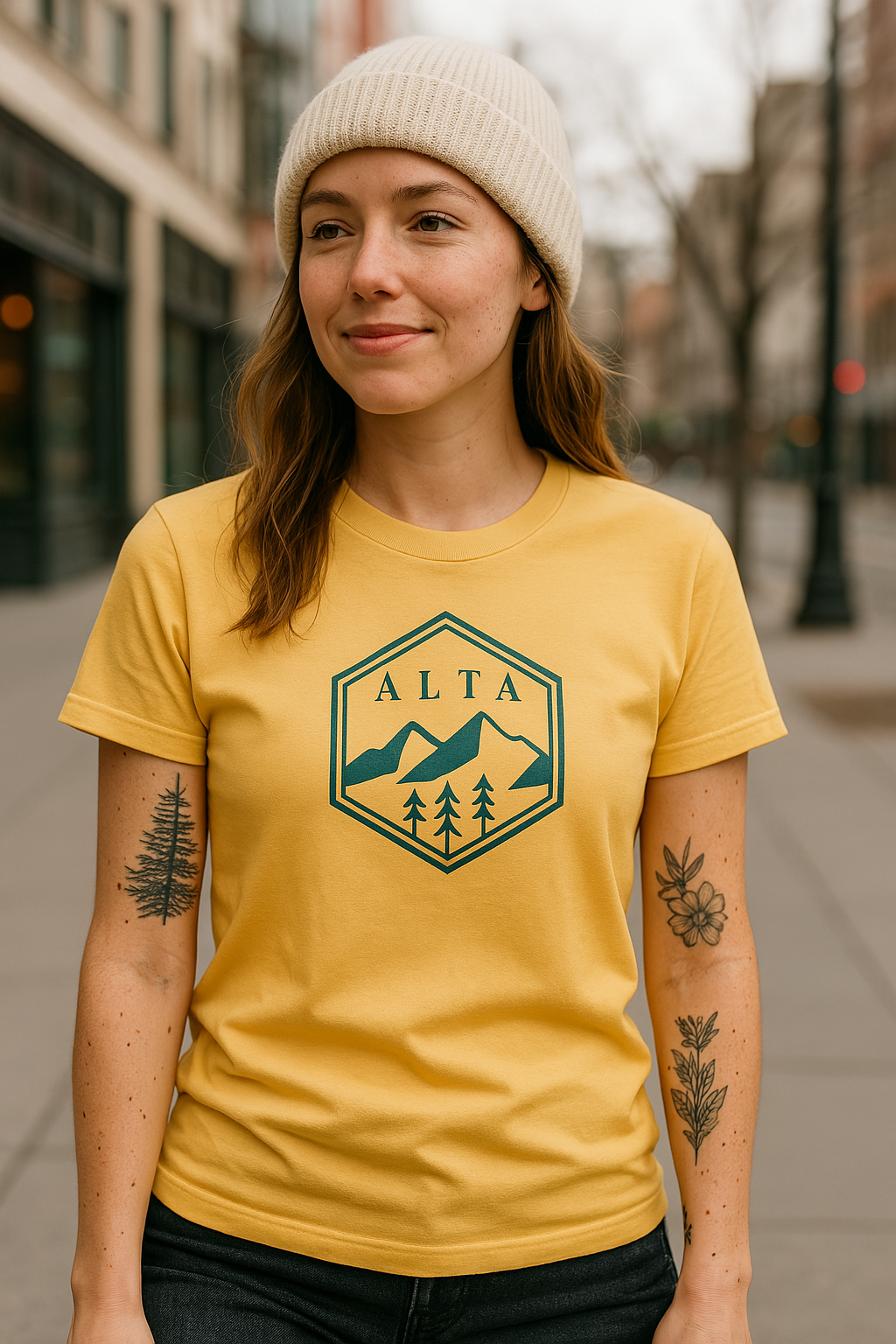 Woman in a mustard yellow t-shirt with a navy "ALTA" mountain and tree emblem design, standing in an urban downtown street setting. She is wearing a cream knit beanie and has visible botanical tattoos on both arms.
