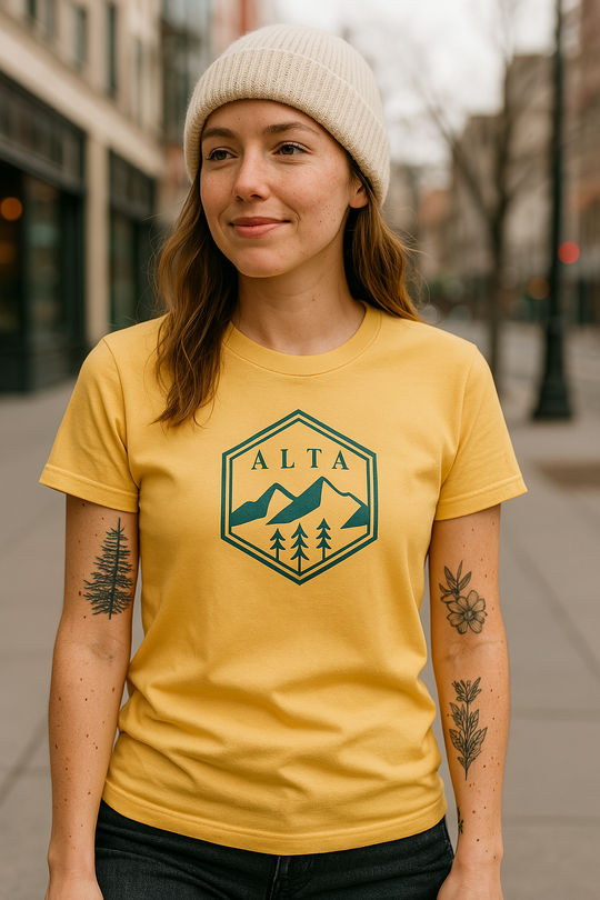 Woman in a mustard yellow t-shirt with a navy "ALTA" mountain and tree emblem design, standing in an urban downtown street setting. She is wearing a cream knit beanie and has visible botanical tattoos on both arms.
