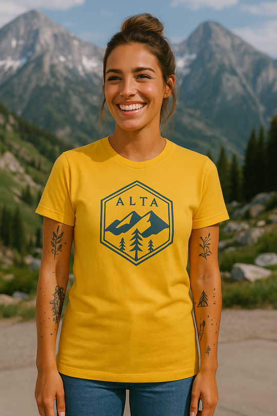 Woman wearing a bright yellow t-shirt with a navy blue hexagon design that features the word "ALTA" above a mountain and pine tree graphic, captured in a scenic alpine setting with rugged peaks and lush greenery in the background.